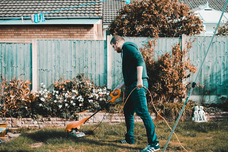 A man with short dark hair and a beard, wearing a checkered shirt, black apron, white gloves, and light-colored jeans, crouches on a well-maintained grassy lawn while operating a black petrol-powered 