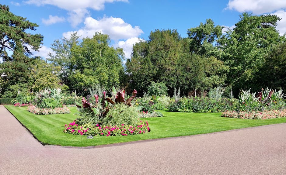 A well-maintained garden featuring lush, vibrant green grass in the foreground, bordered by a narrow pathway with freshly turned dark soil along its edges. To the left and right, there are neatly trim